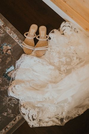 Elegant white lace wedding dress with sandals laid out on dark surface.