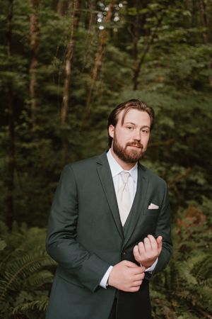 A groom in a dark suit adjusts his cuff in a forest setting.