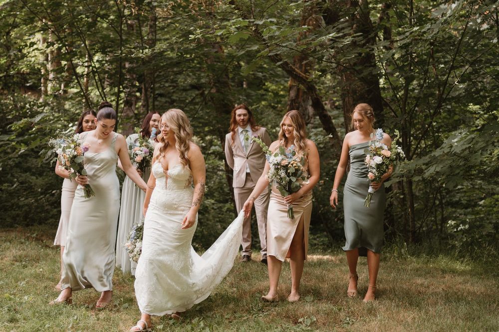 Wedding party in white dresses walks through a sunlit forest path.