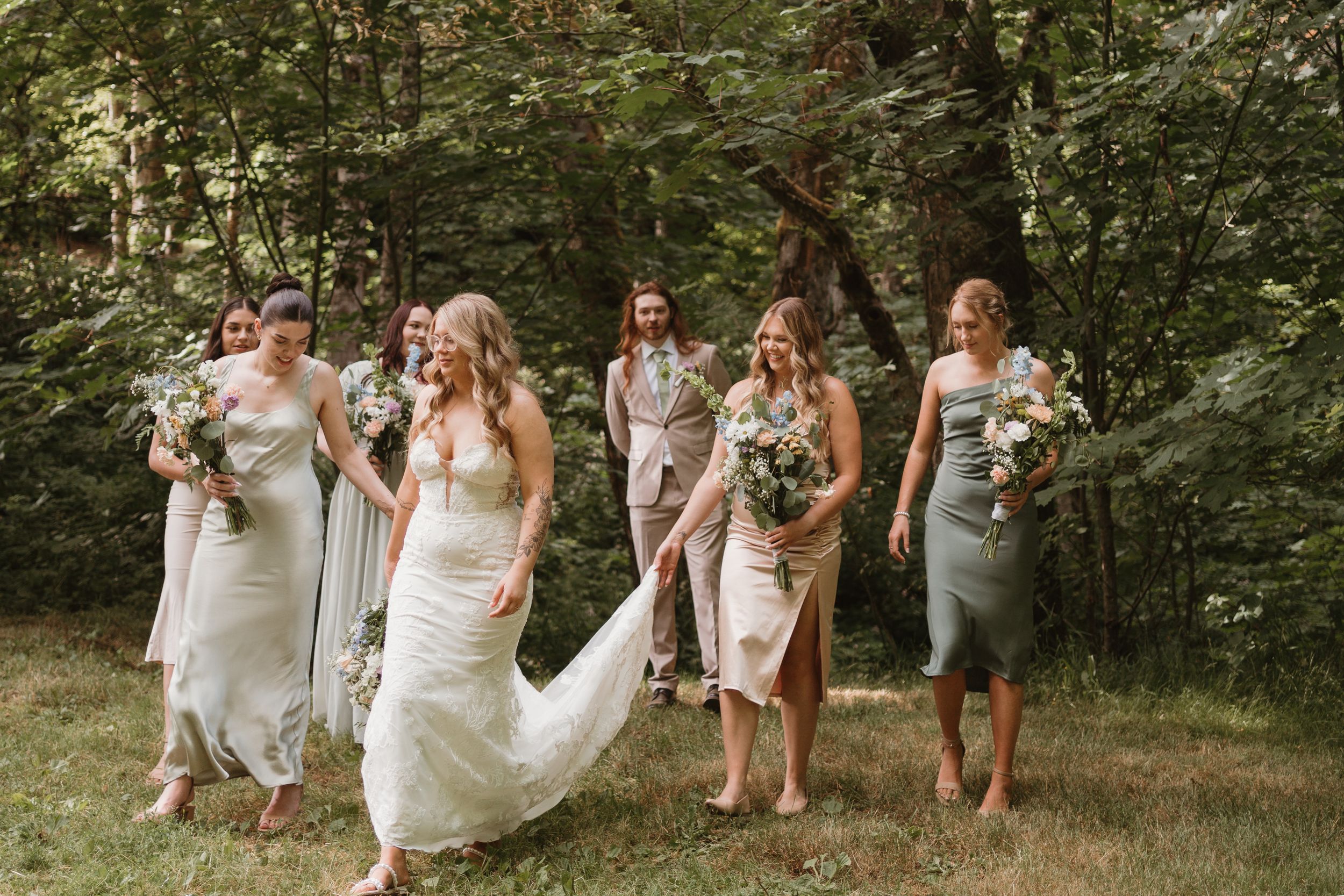 Wedding party in white dresses walks through a sunlit forest path.