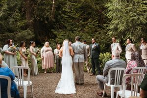 Outdoor forest wedding ceremony with guests seated on wooden chairs among tall trees.