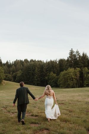Newlyweds walk hand in hand through a scenic hillside meadow bordered by evergreen forest.