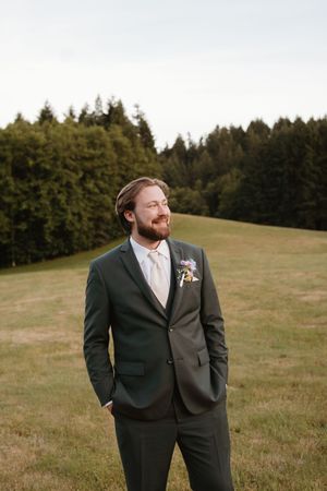 A groom in a dark suit poses joyfully on a grassy hillside with evergreen trees in the background.