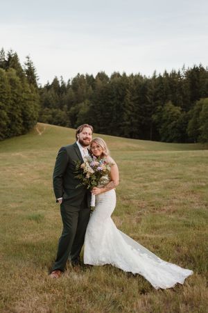 A wedding couple poses together in a meadow with a beautiful forest backdrop.