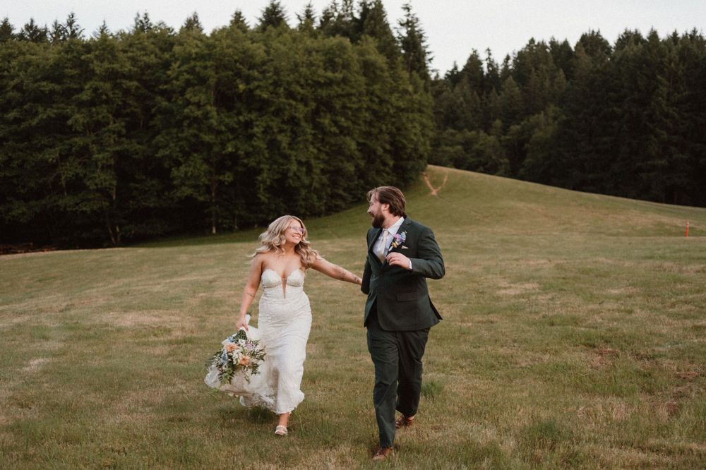 Playful wedding couple runs through a scenic meadow surrounded by evergreen trees.