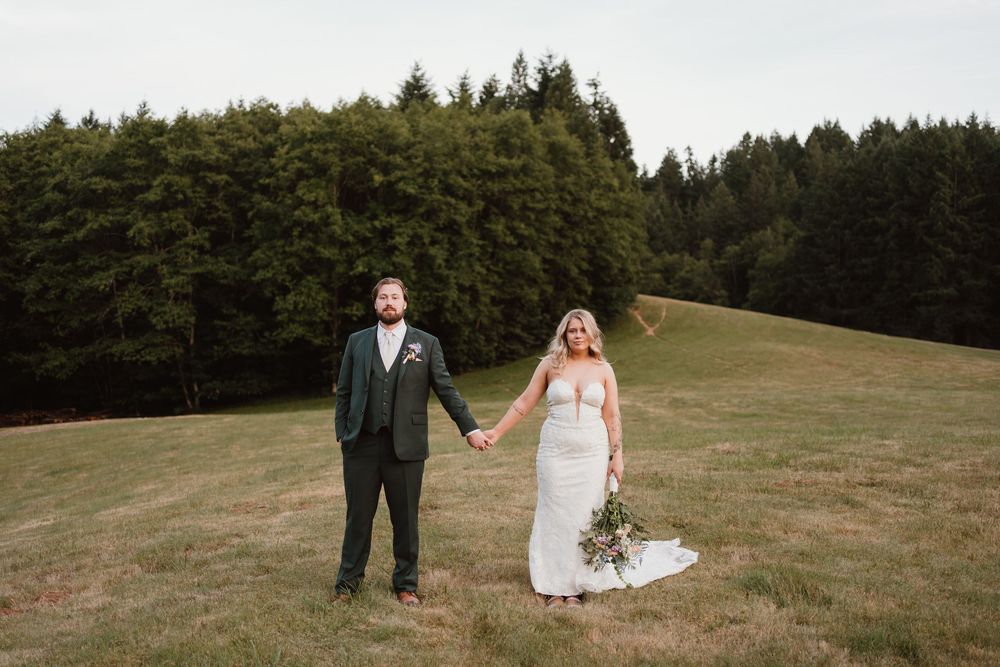 Wedding couple poses together on a grassy hillside with evergreen forest in the background.