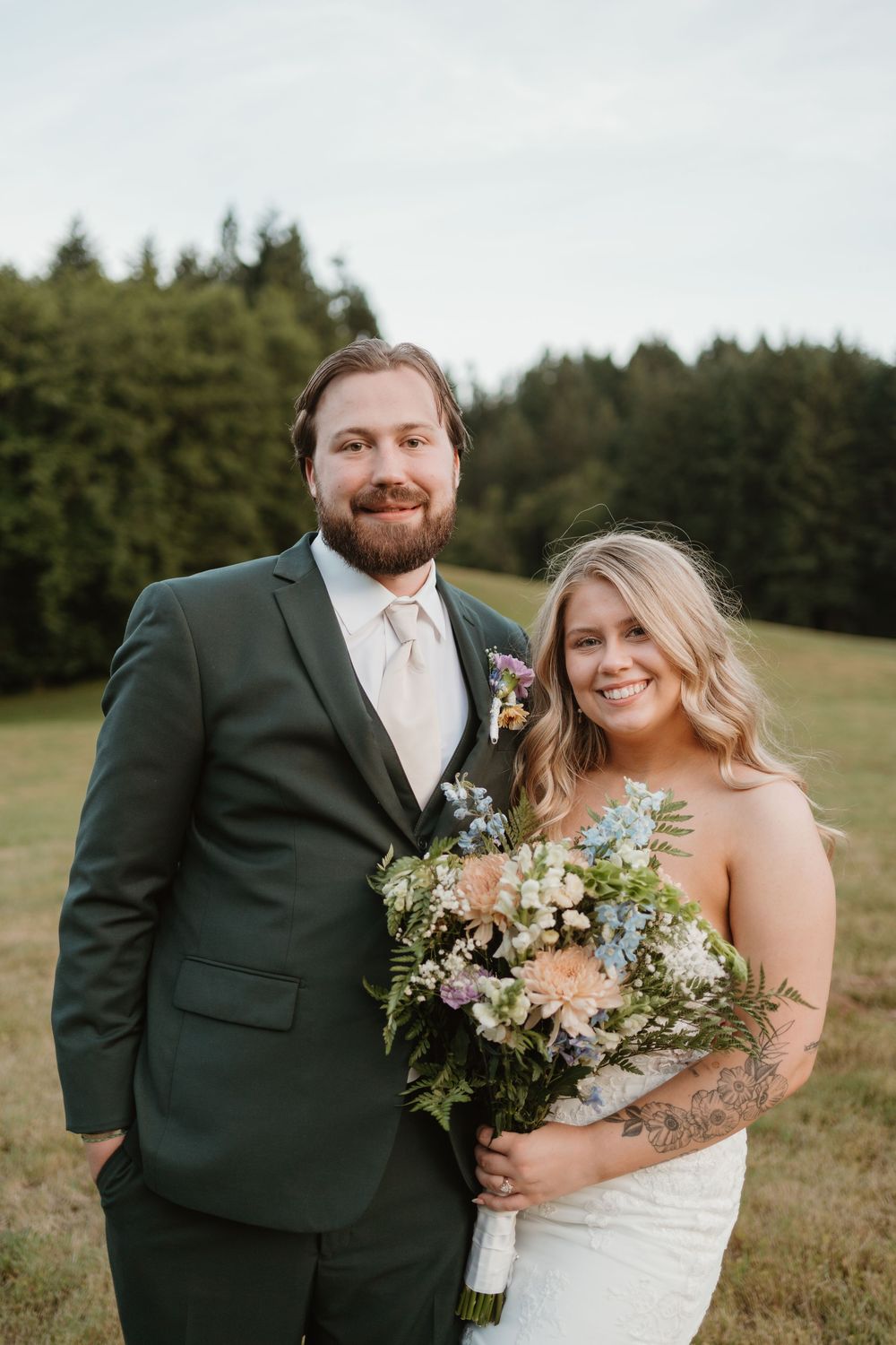 Wedding couple poses in a grassy field with forest backdrop and beautiful floral bouquet.