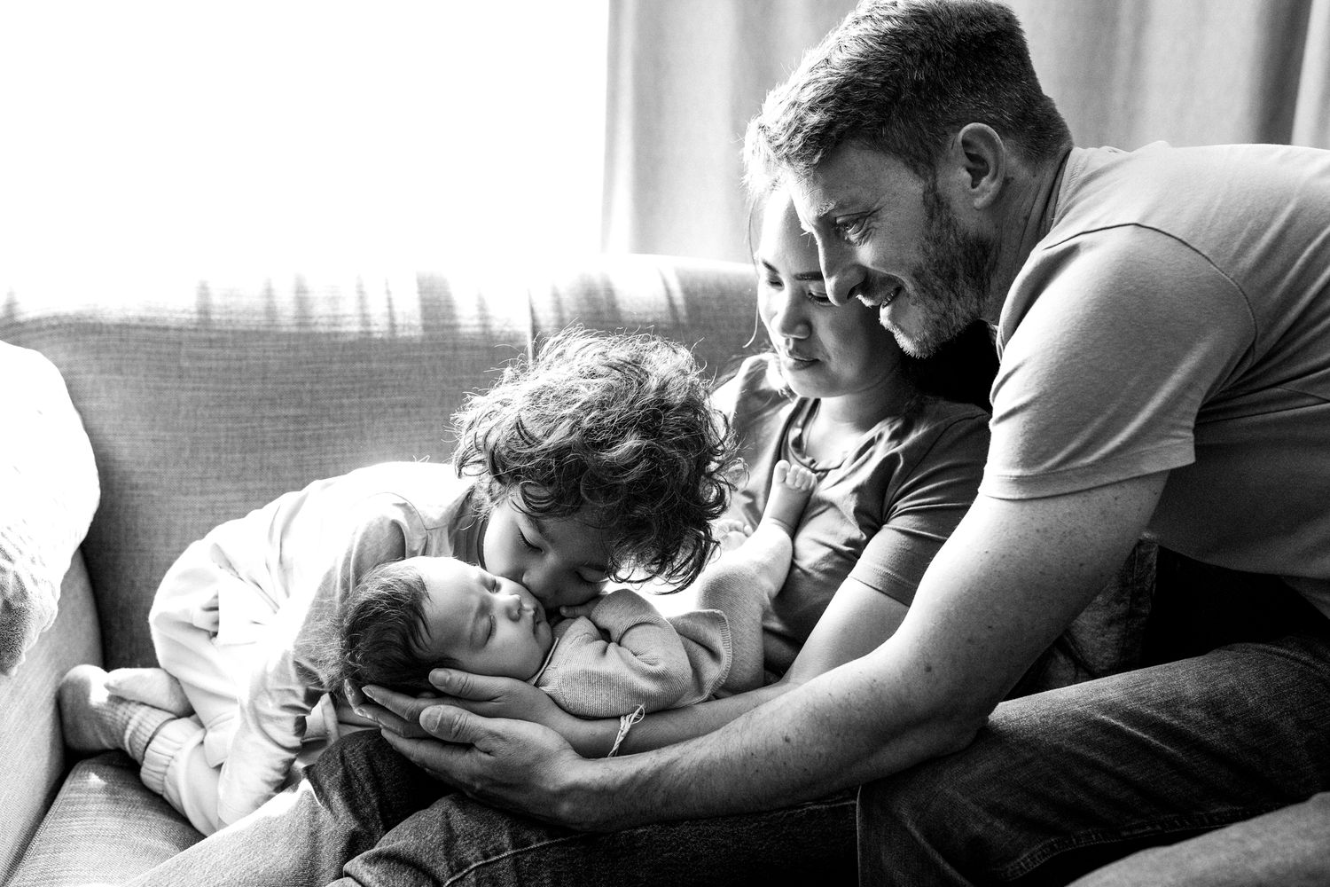 A tender black and white moment captured on a couch as a family looks down at a baby together.