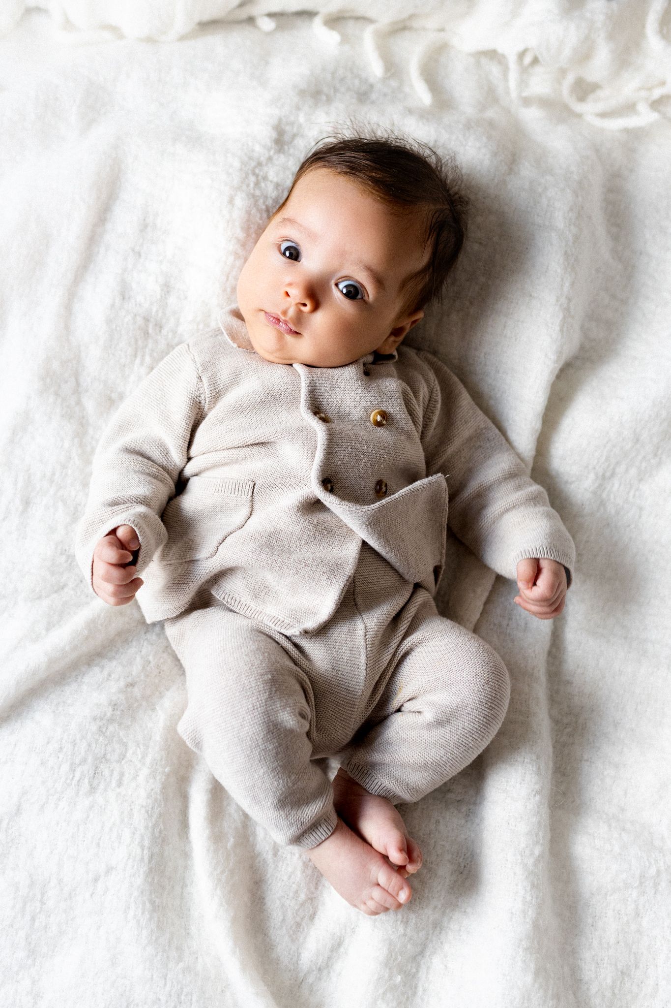A small baby wearing a cozy beige onesie lies on soft white bedding while looking directly at the camera.