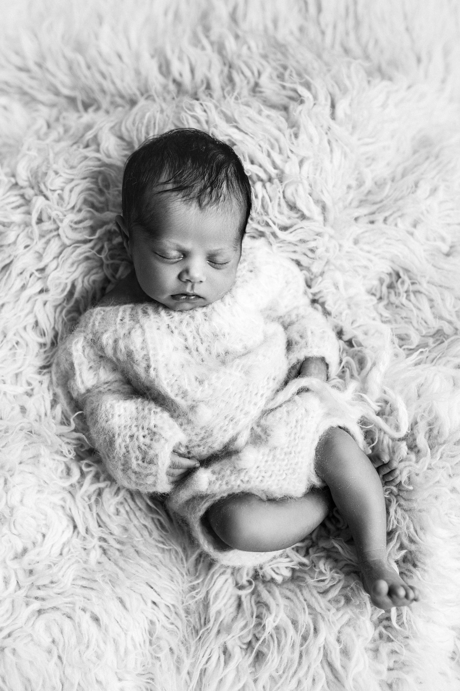 A sleeping baby wrapped in white lace blanket lies peacefully on soft white fur rug in black and white photograph.
