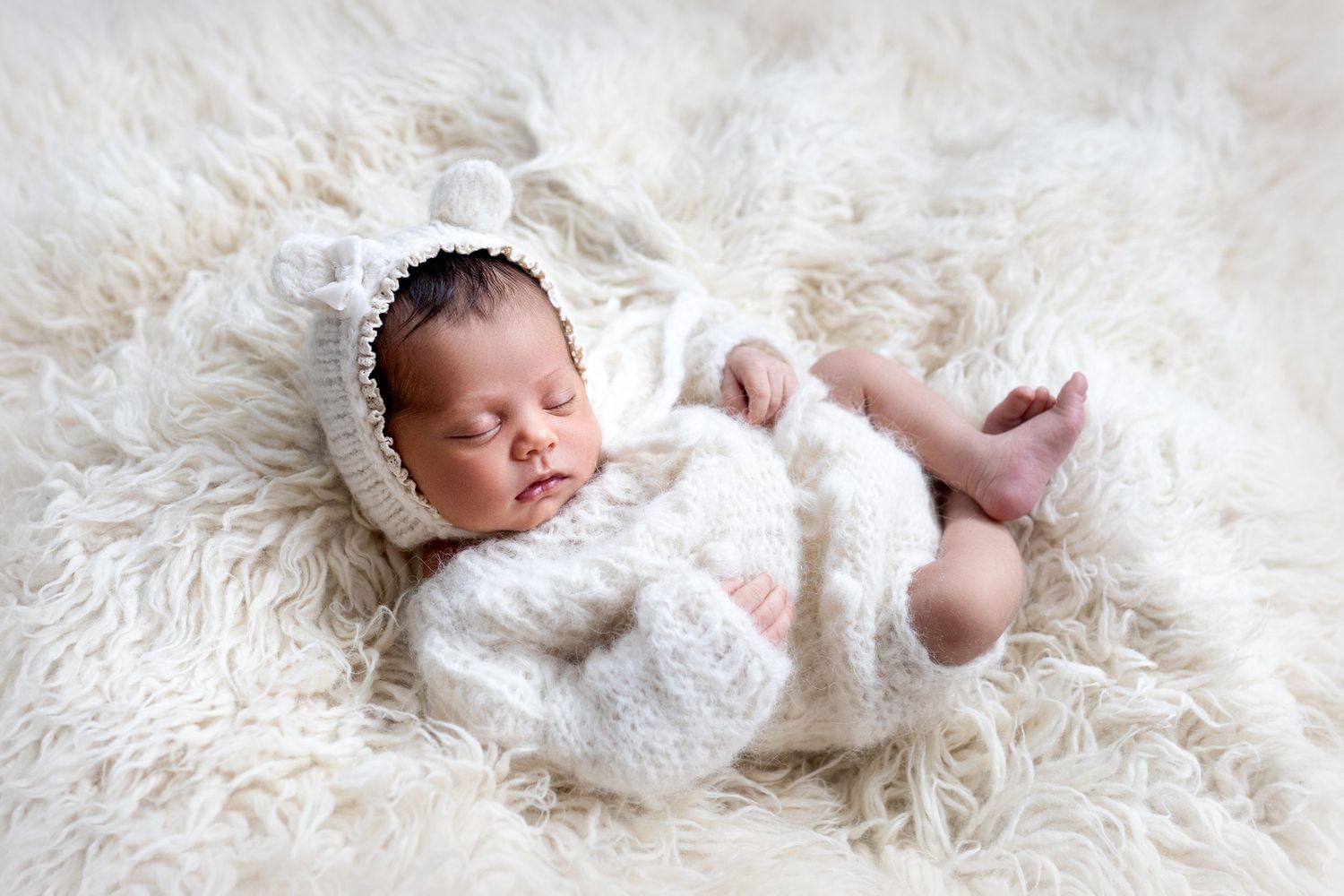 A newborn baby sleeps peacefully on soft white fur during a professional photo session.