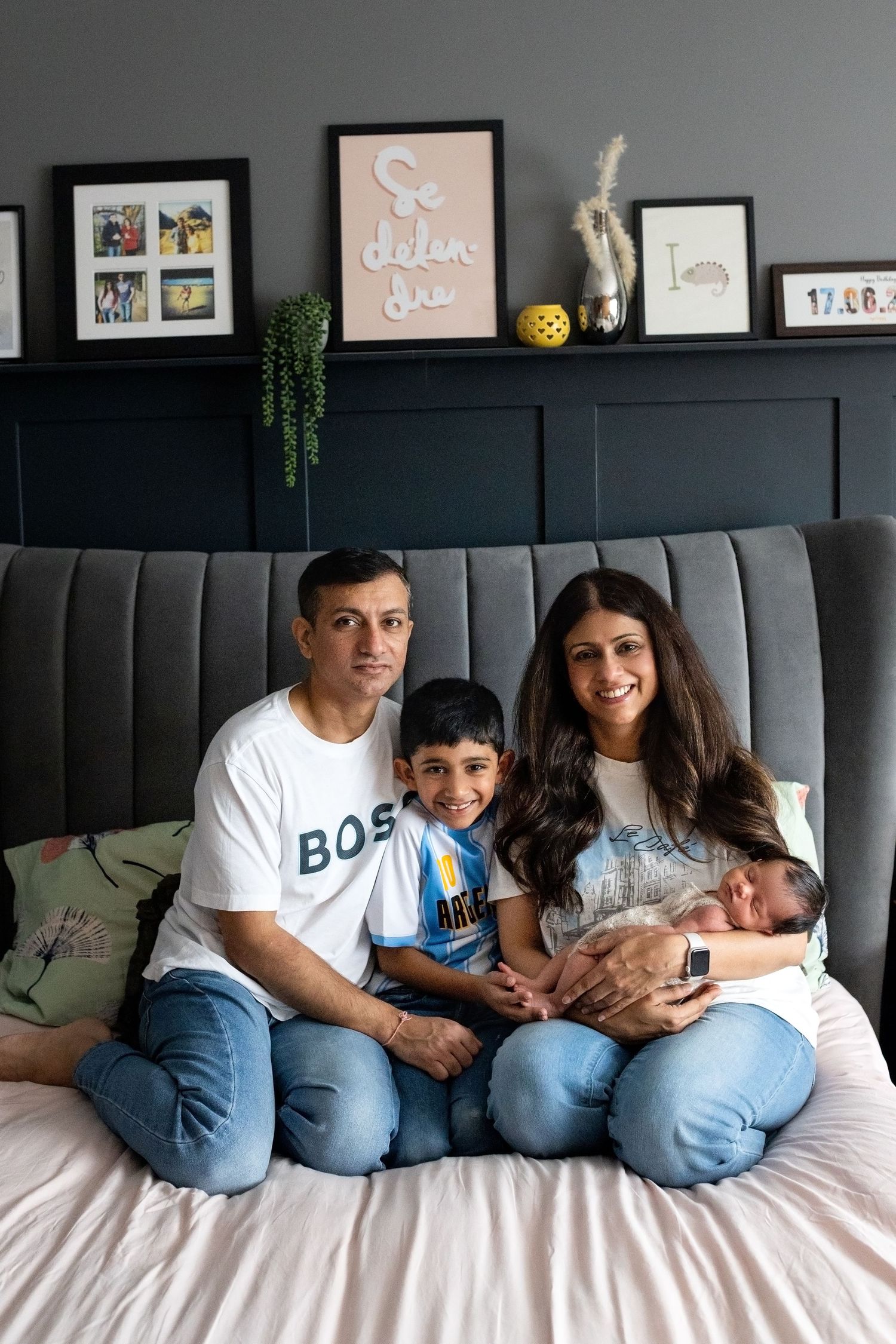 A family lounges together on a gray couch with decorative frames displayed on the dark wall behind them.