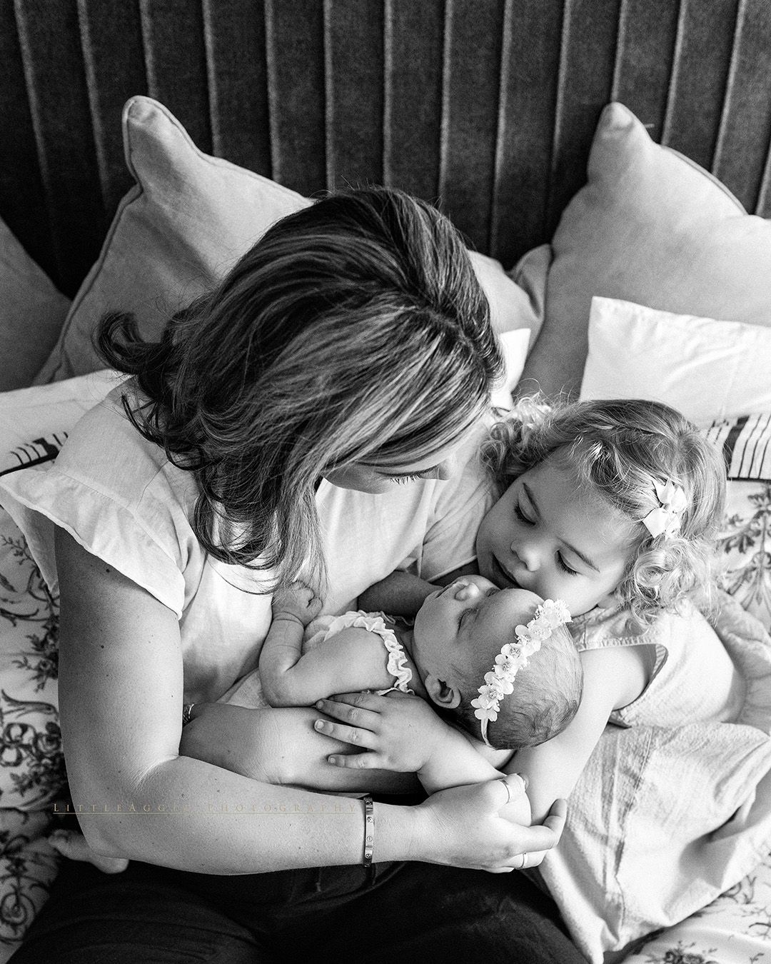Black and white intimate portrait of family members sharing tender moment while resting together on bed with pillows.