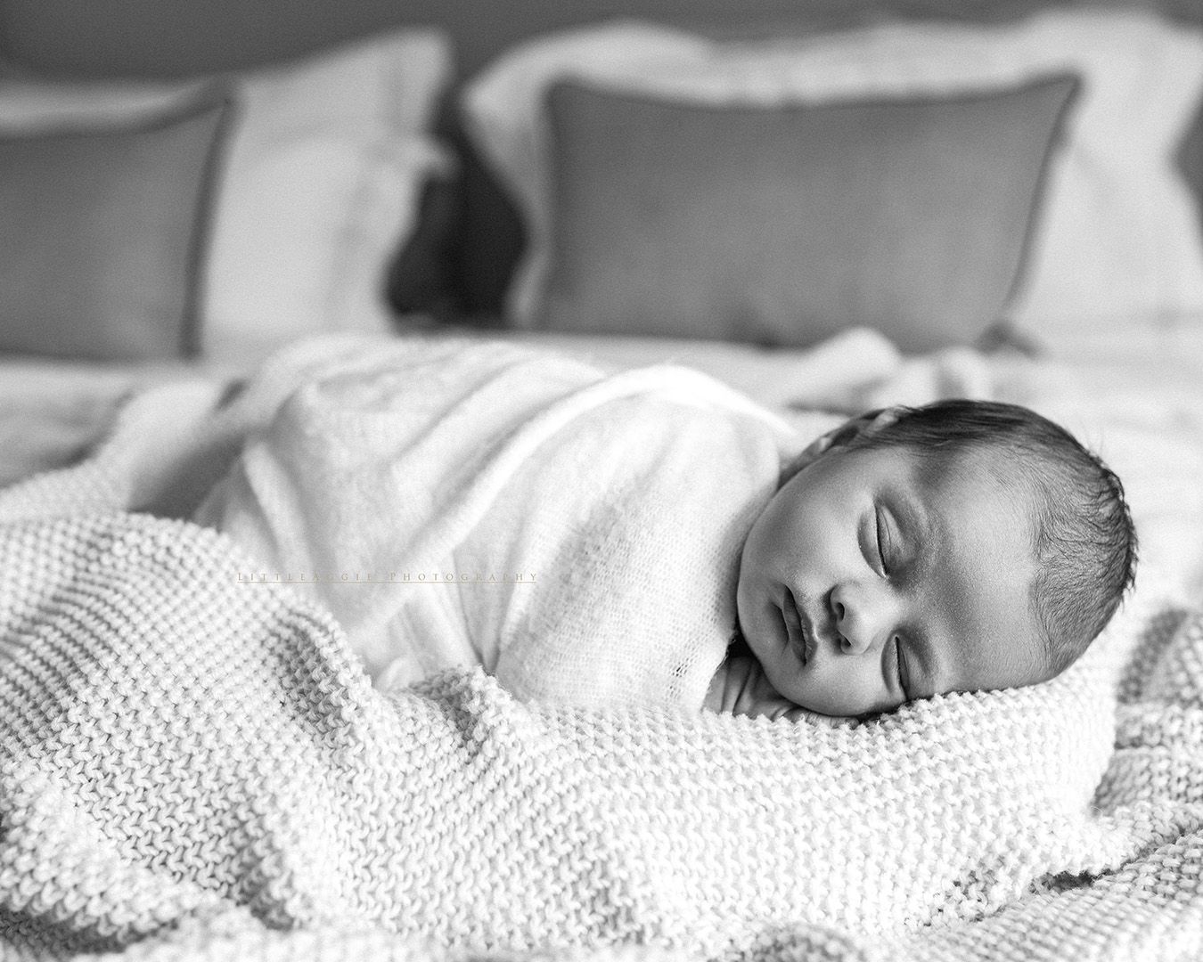 A peaceful newborn baby wrapped in a cozy white knit blanket sleeping on soft pillows, captured in black and white.