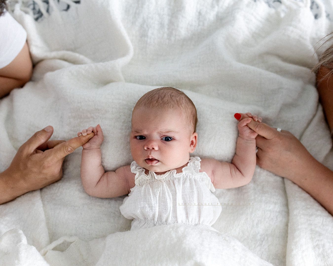 A small baby in a white outfit lays on white bedding while adult hands gently touch their arms.