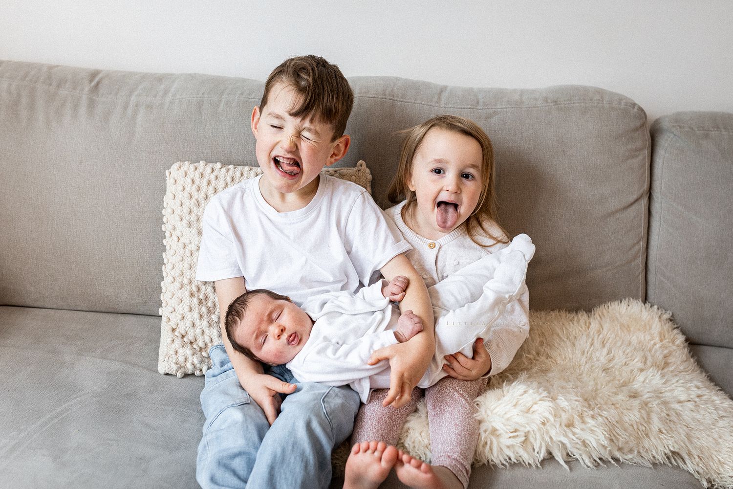 Children in white clothing playing together on a gray couch with plush pillows and blankets.
