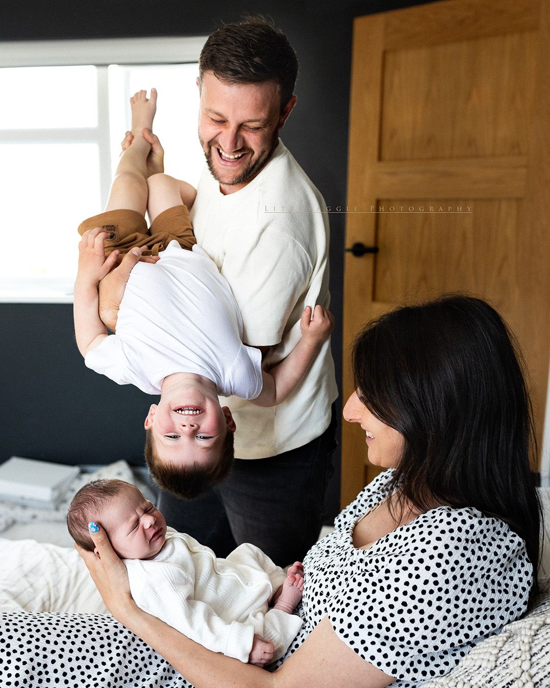 A joyful family moment captured on a bed as a parent holds a child upside down while another parent holds a baby.