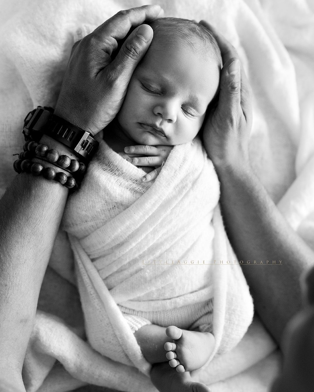 Close-up black and white photograph of a sleeping newborn baby swaddled in a soft white blanket.