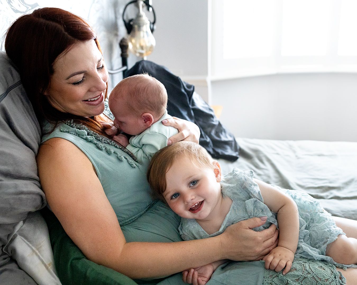 A tender family moment with mother and two small children snuggling on white bedding while relaxing at home.