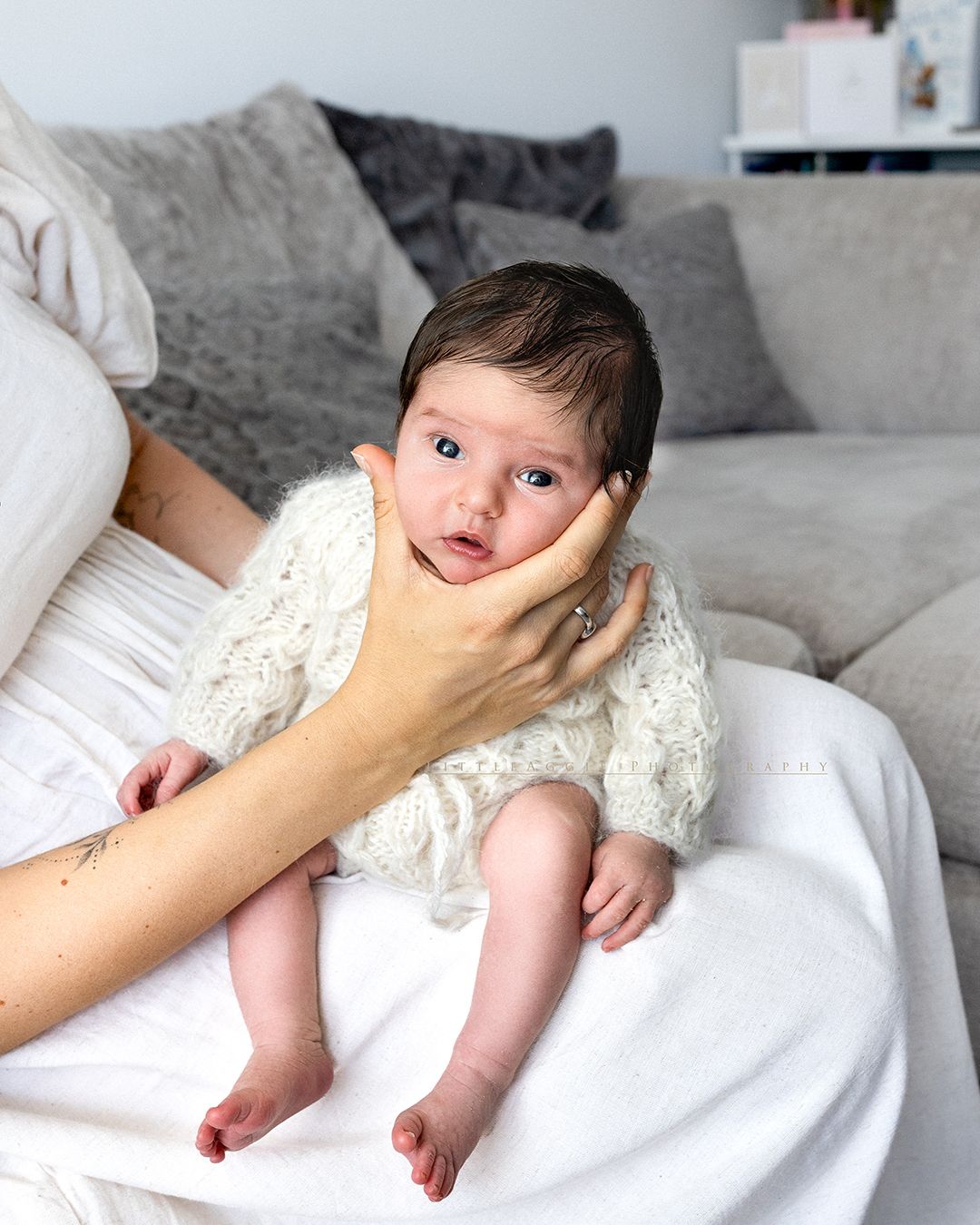 A newborn baby in a white knit outfit being cradled by a mother's hand on a soft white blanket.