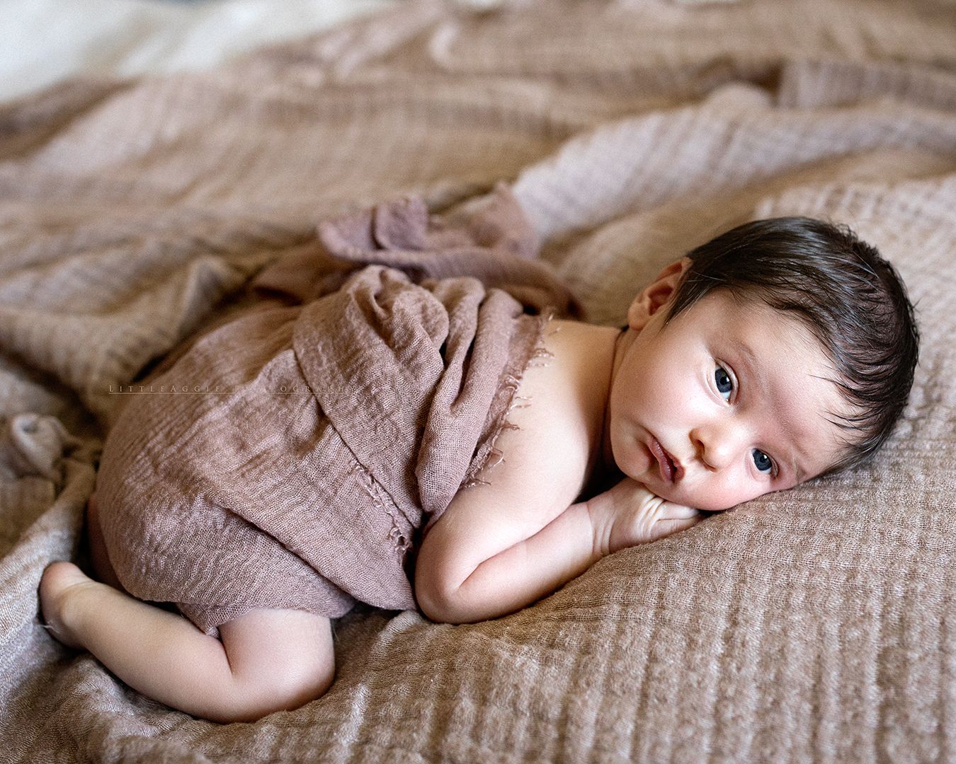 Newborn baby wrapped in soft mauve muslin cloth, lying on a beige knit blanket in a peaceful pose.