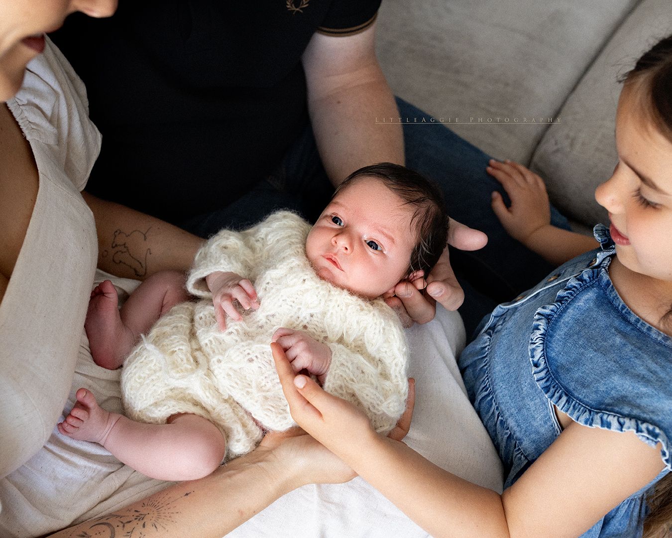 A newborn baby in a white knit outfit being held by a mother while an older sibling looks on lovingly.