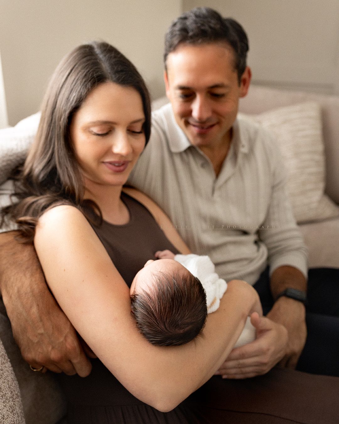 New parents smiling and gazing lovingly at their newborn baby in a warm, cozy indoor setting.