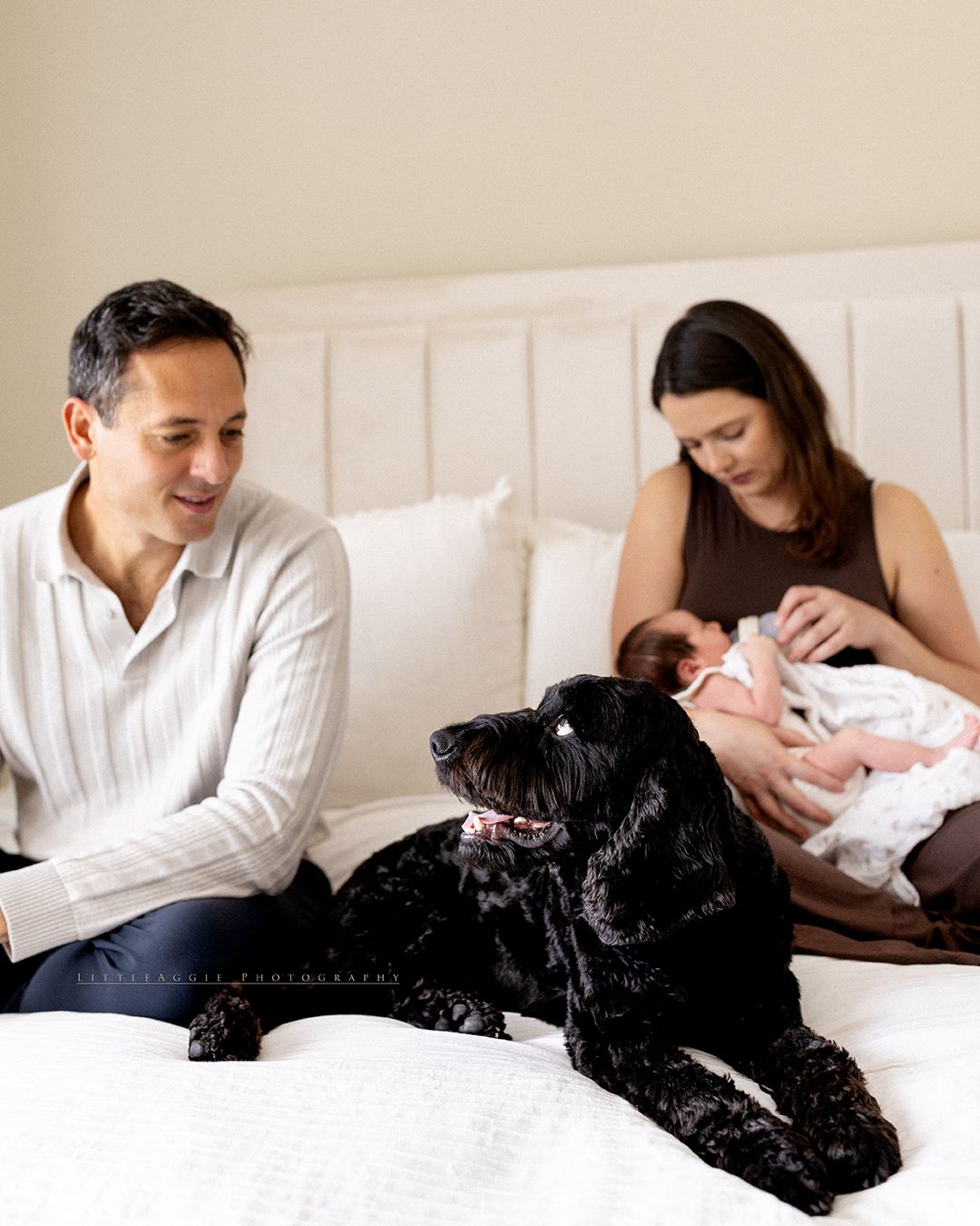 Family with newborn baby and black dog relaxing on bed in a warm, intimate home setting.