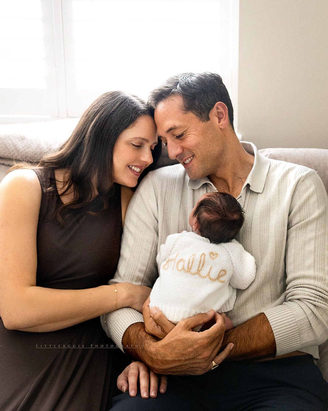 New parents smiling lovingly at their newborn baby named Hallie, wrapped in a white personalized blanket.