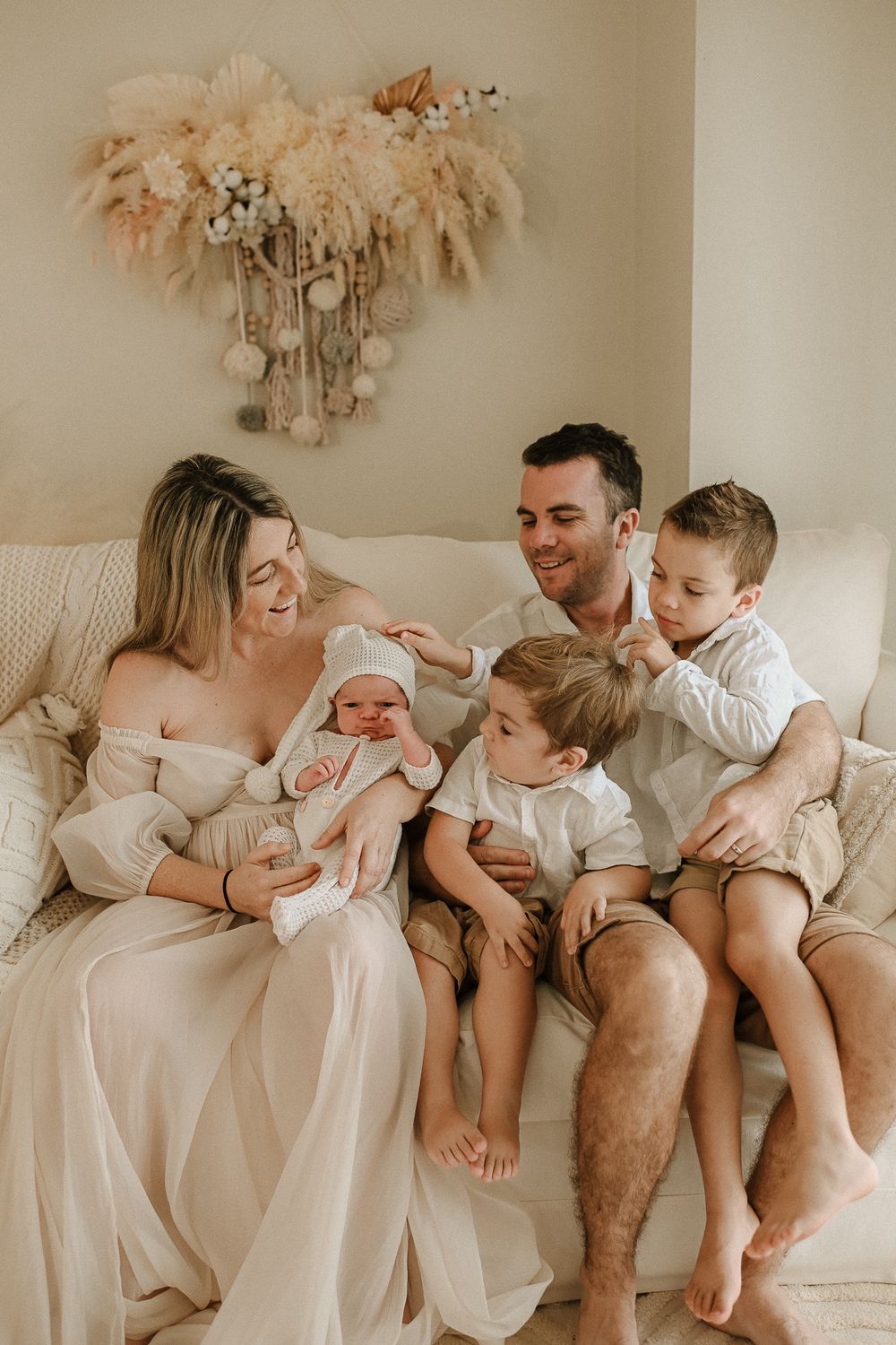 A family shares an intimate moment on a cream couch beneath a decorative pampas grass wall hanging.