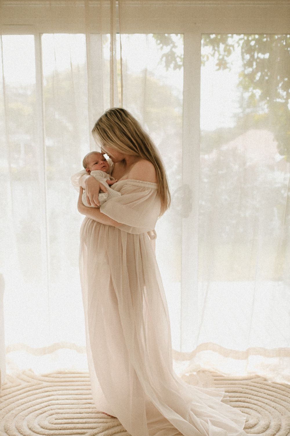 A silhouetted figure in a flowing dress holds a baby against sunlit windows in a newborn studio located in southern Sydney.