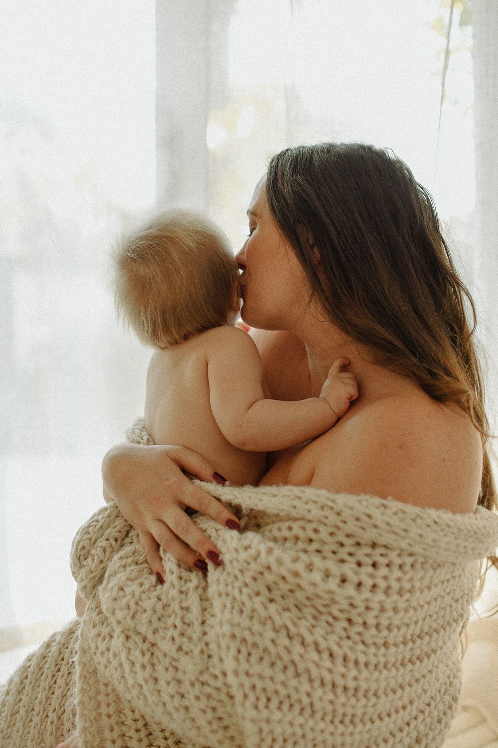 Parent in a knit blanket holding and kissing a baby near a window with soft natural light.