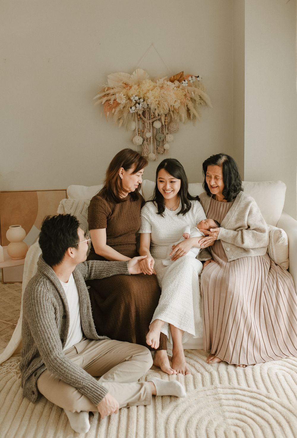 Group sitting on white couch under dried pampas grass decoration in neutral toned room.