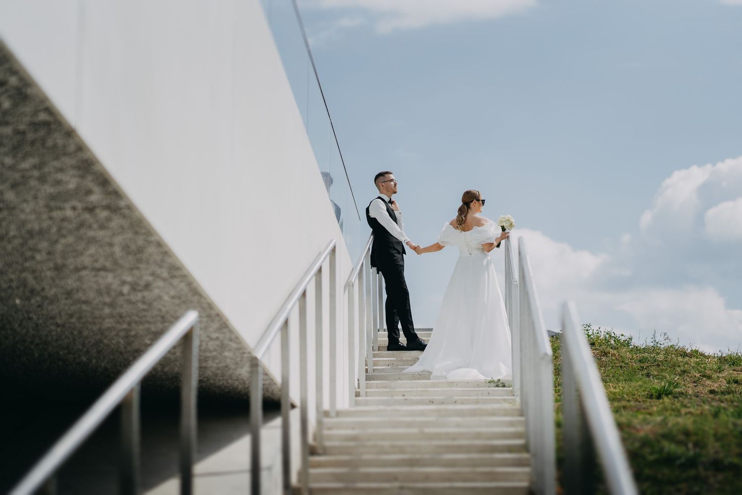 Wedding couple ascends white staircase against bright blue sky.