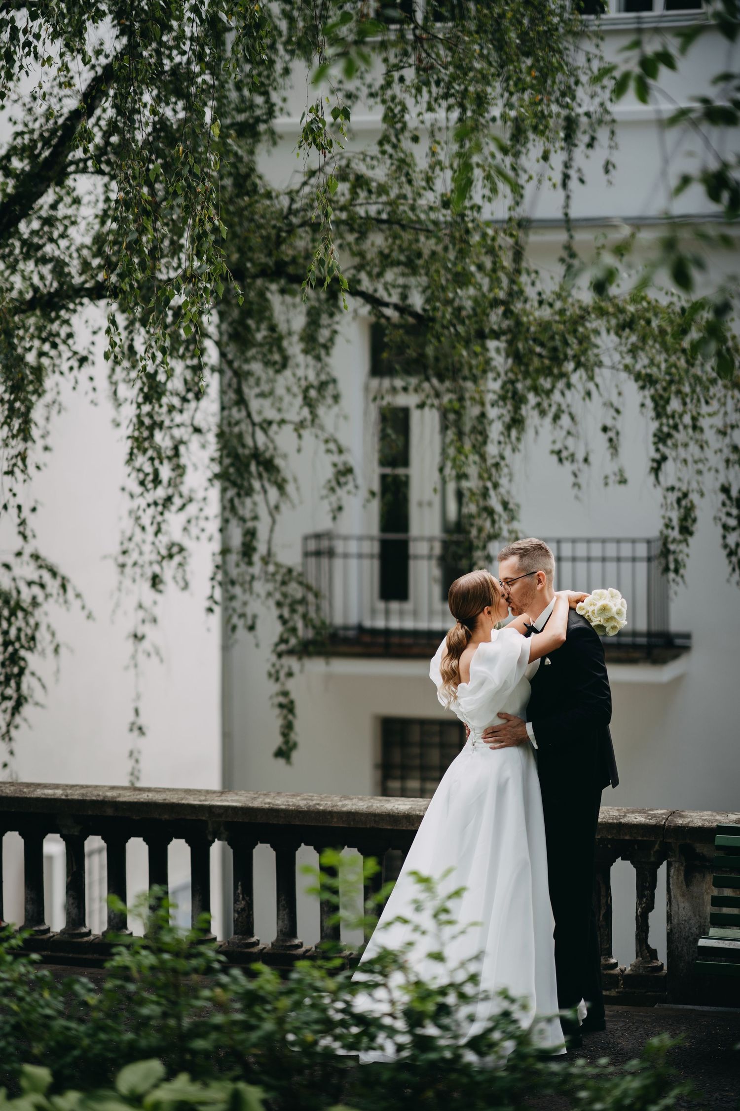Elegant couple dances beneath drooping birch trees against white building facade.