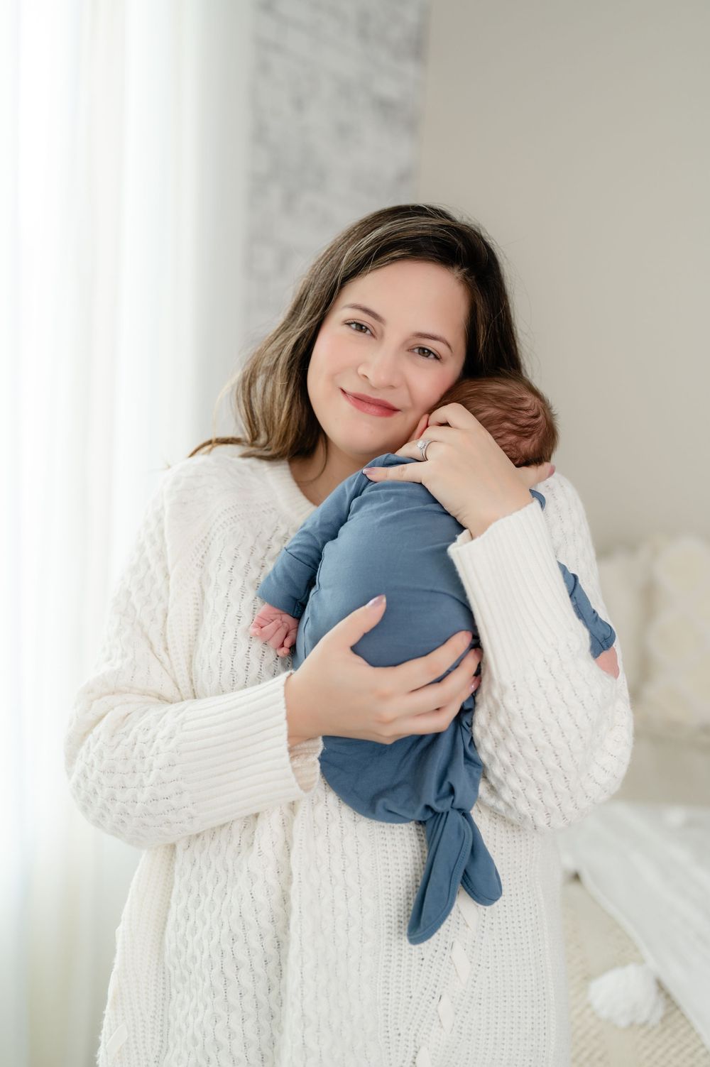 A parent wearing a cozy white cable knit sweater cradles and snuggles with a sleeping baby in a blue outfit.