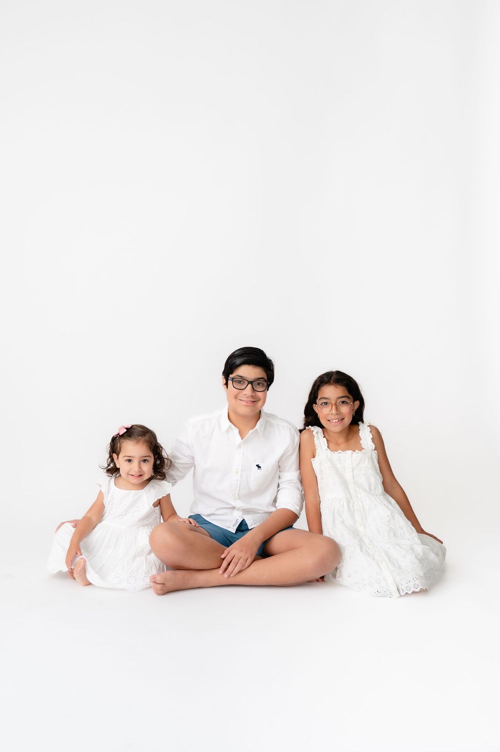 Three children in white clothing sitting together against a white background for a minimalist studio portrait.