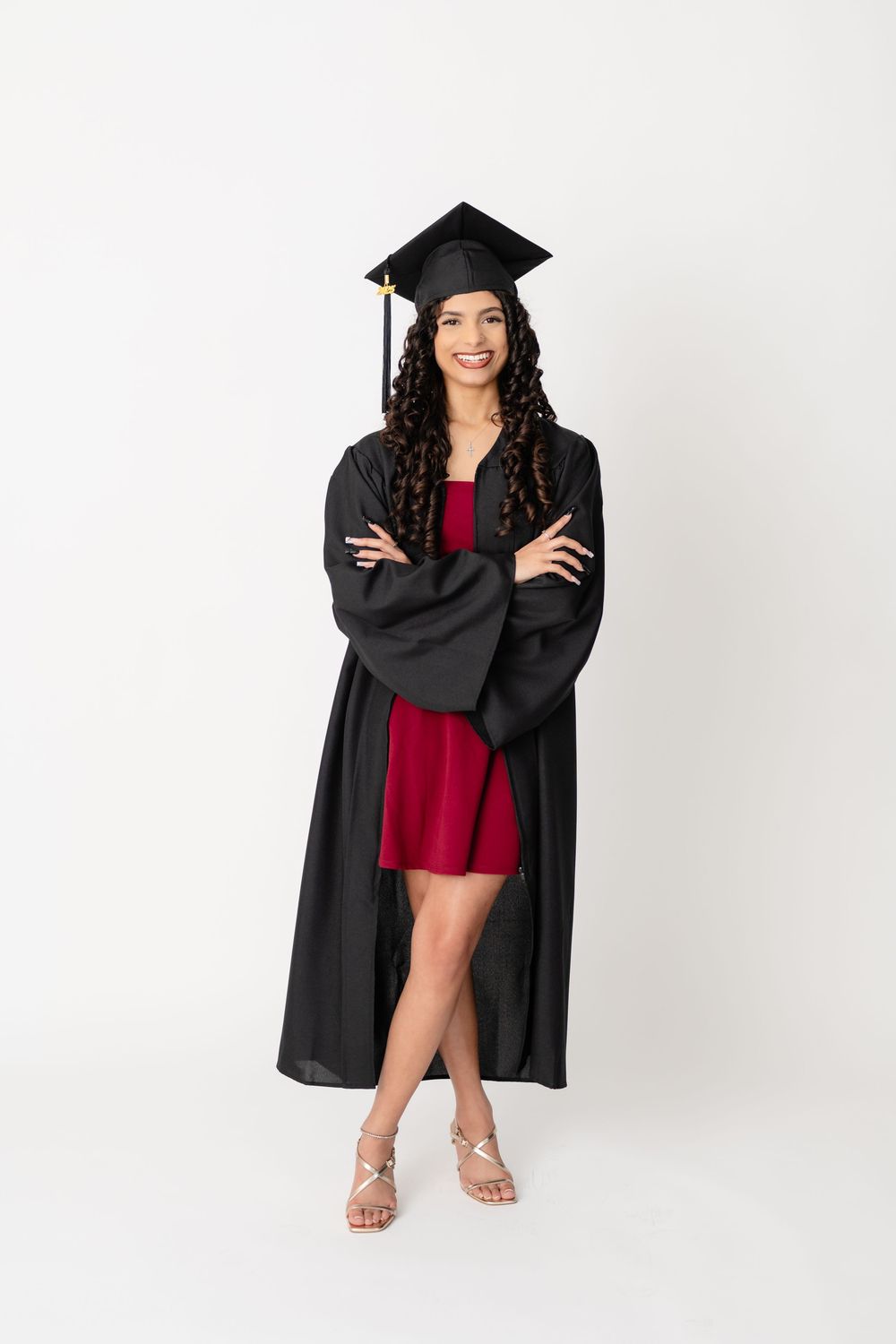 Graduate in black cap and gown and red dress poses confidently with crossed arms on white background.