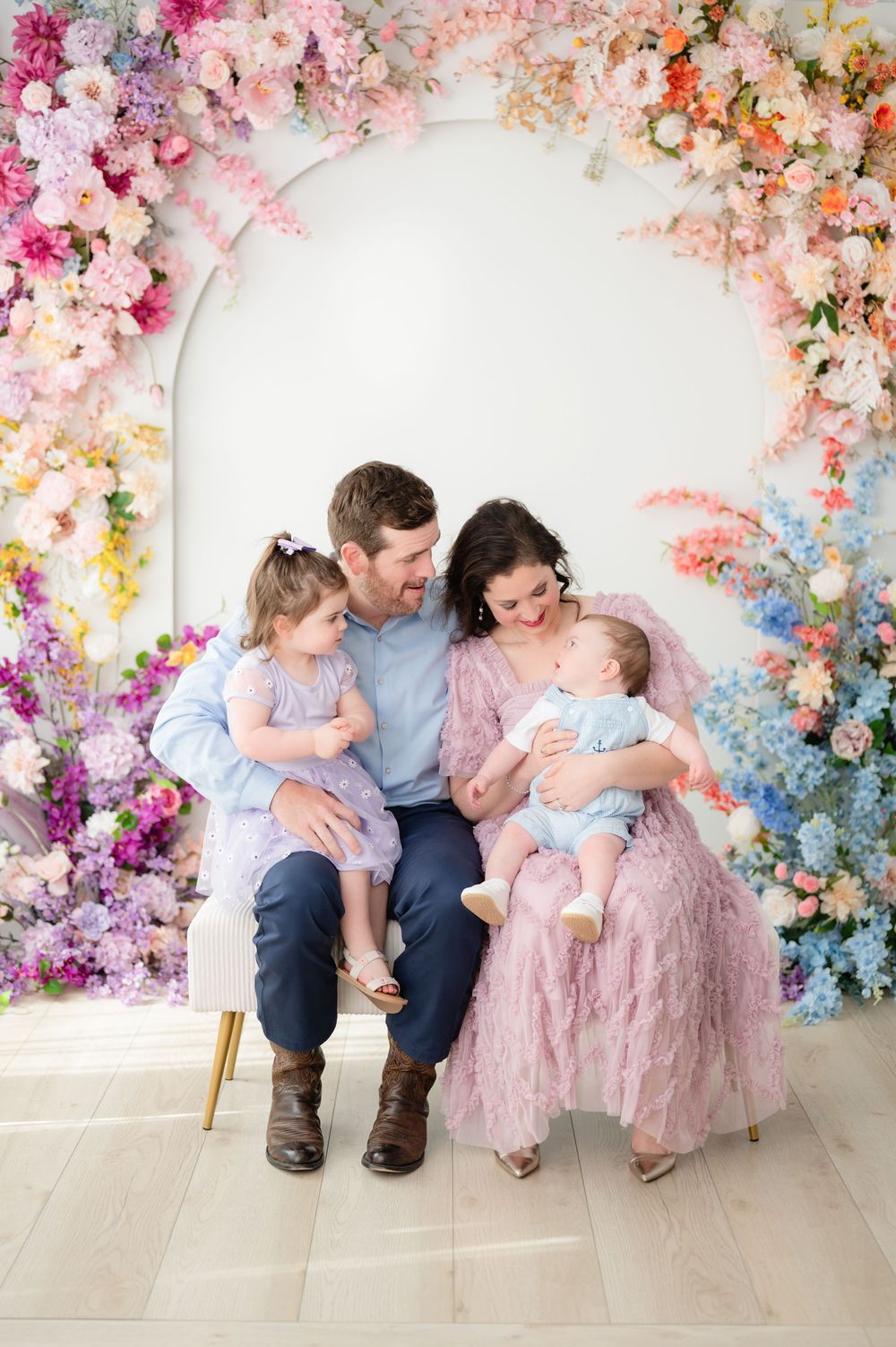 Family sits together on light wood floor against vibrant floral backdrop in pastel pink and blue tones.