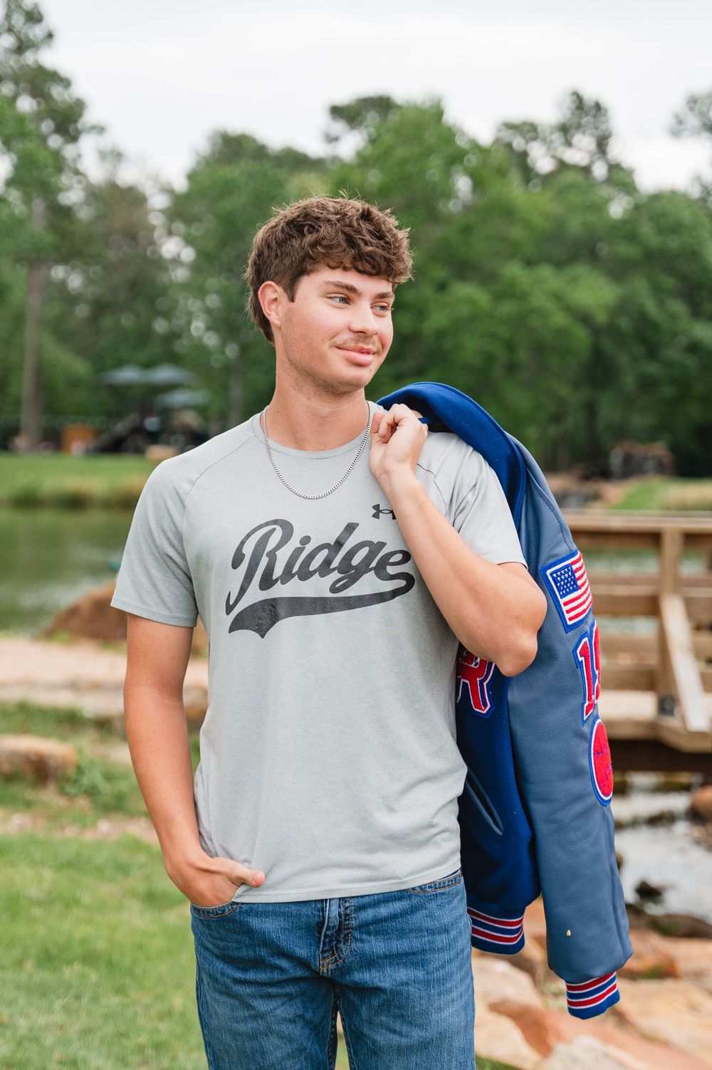 Student wearing a gray Ridge t-shirt holds a blue varsity jacket while standing by a lake with trees in background.