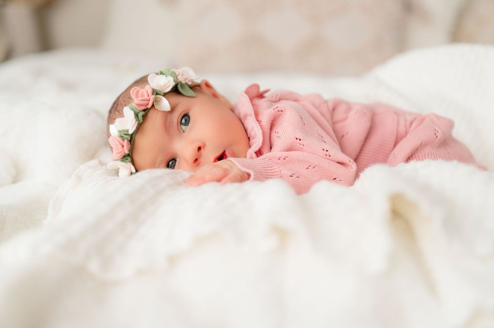 Baby lying on white bedding wearing a pink sweater and delicate floral headband while looking at the camera.