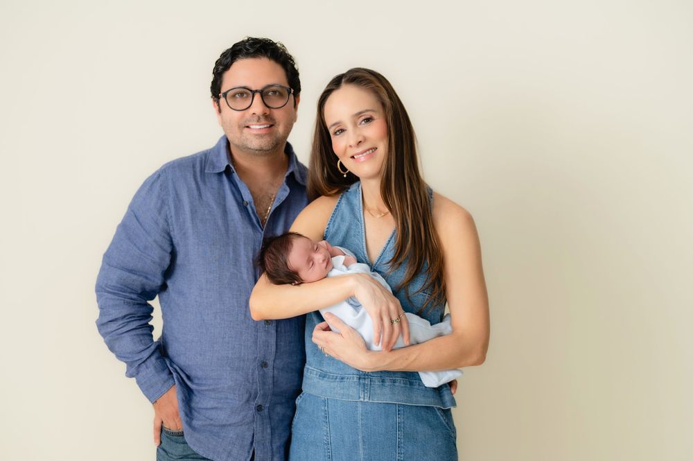 A couple stands together while holding their newborn baby against a light background.