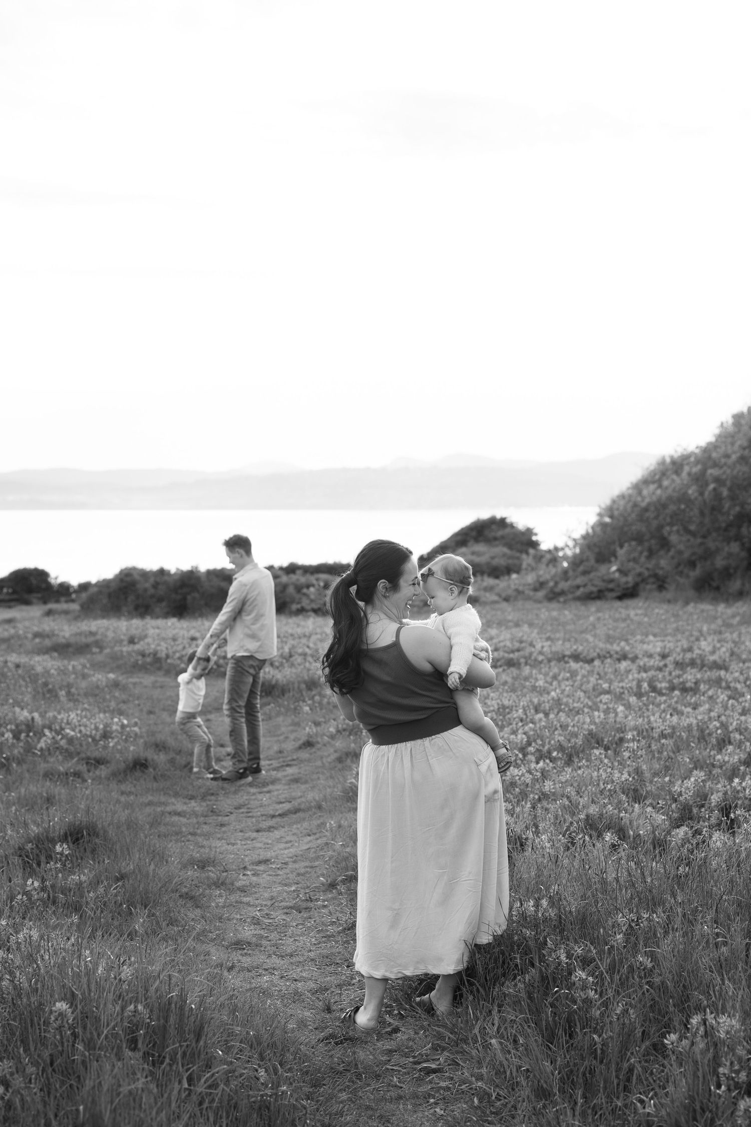 Black and white photo of a mother holding her baby on a coastal meadow path while father walks with toddler in background. Victoria BC.