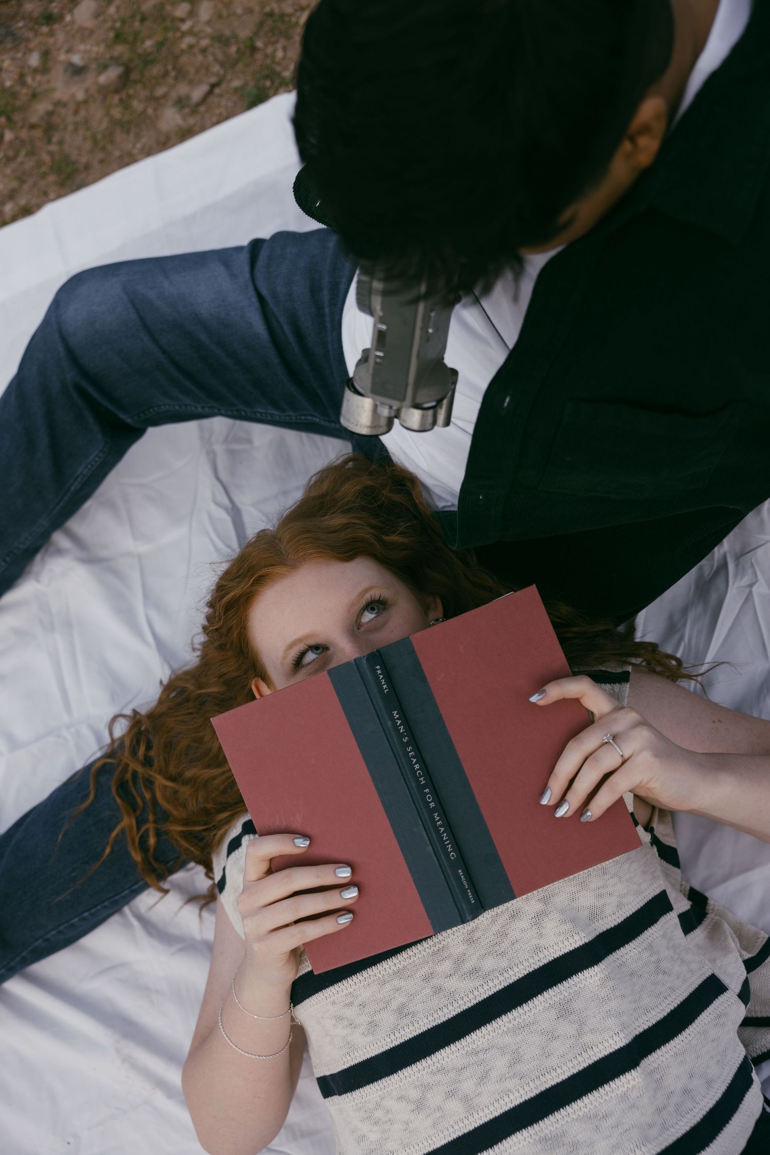 Two people lying on a blanket sharing a red book together outdoors.