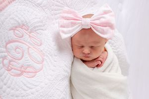 Newborn baby wrapped in white swaddle with pink bow headband resting on textured white blanket.
