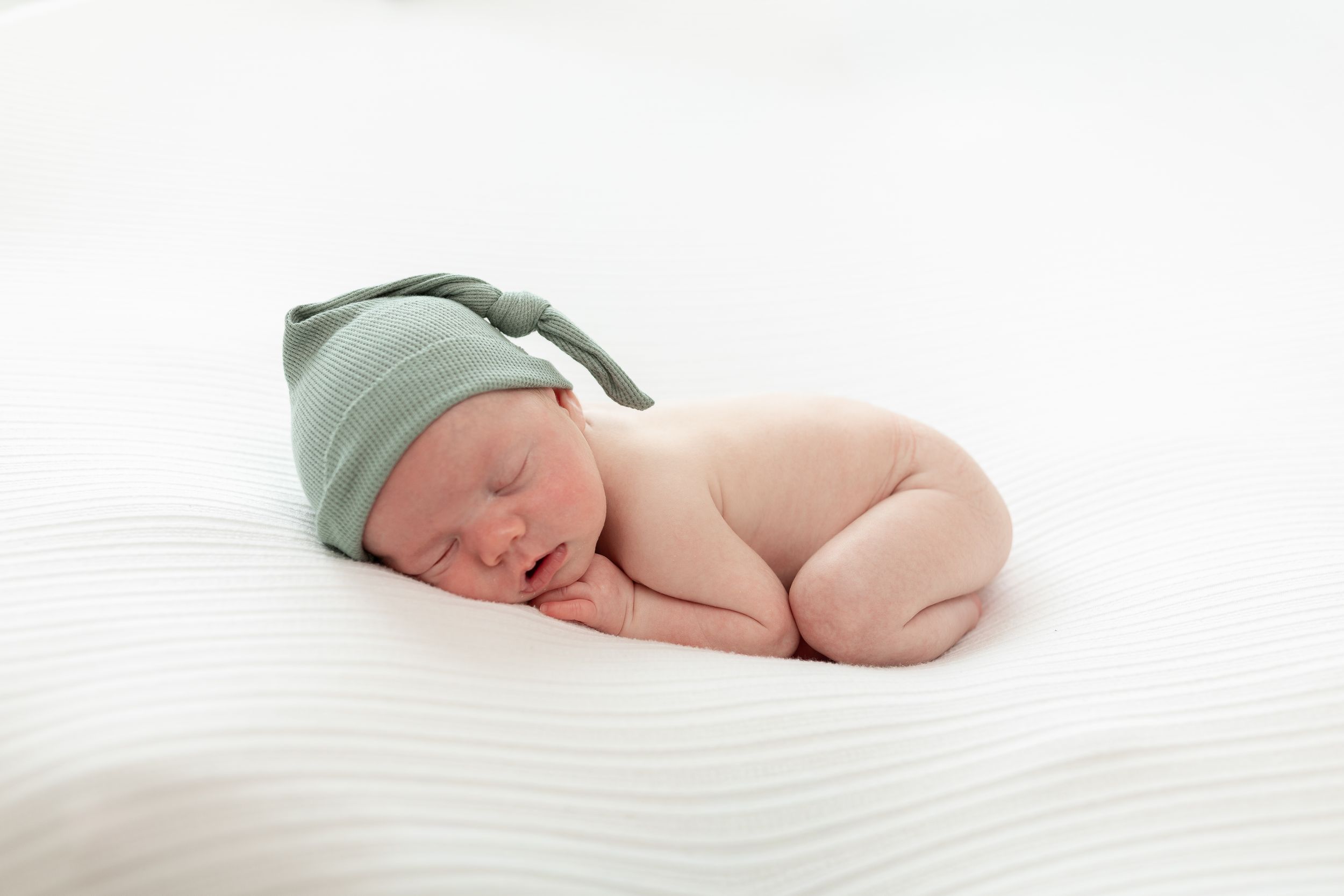 Sleeping newborn baby wearing a green knotted hat, curled up on a white textured blanket.