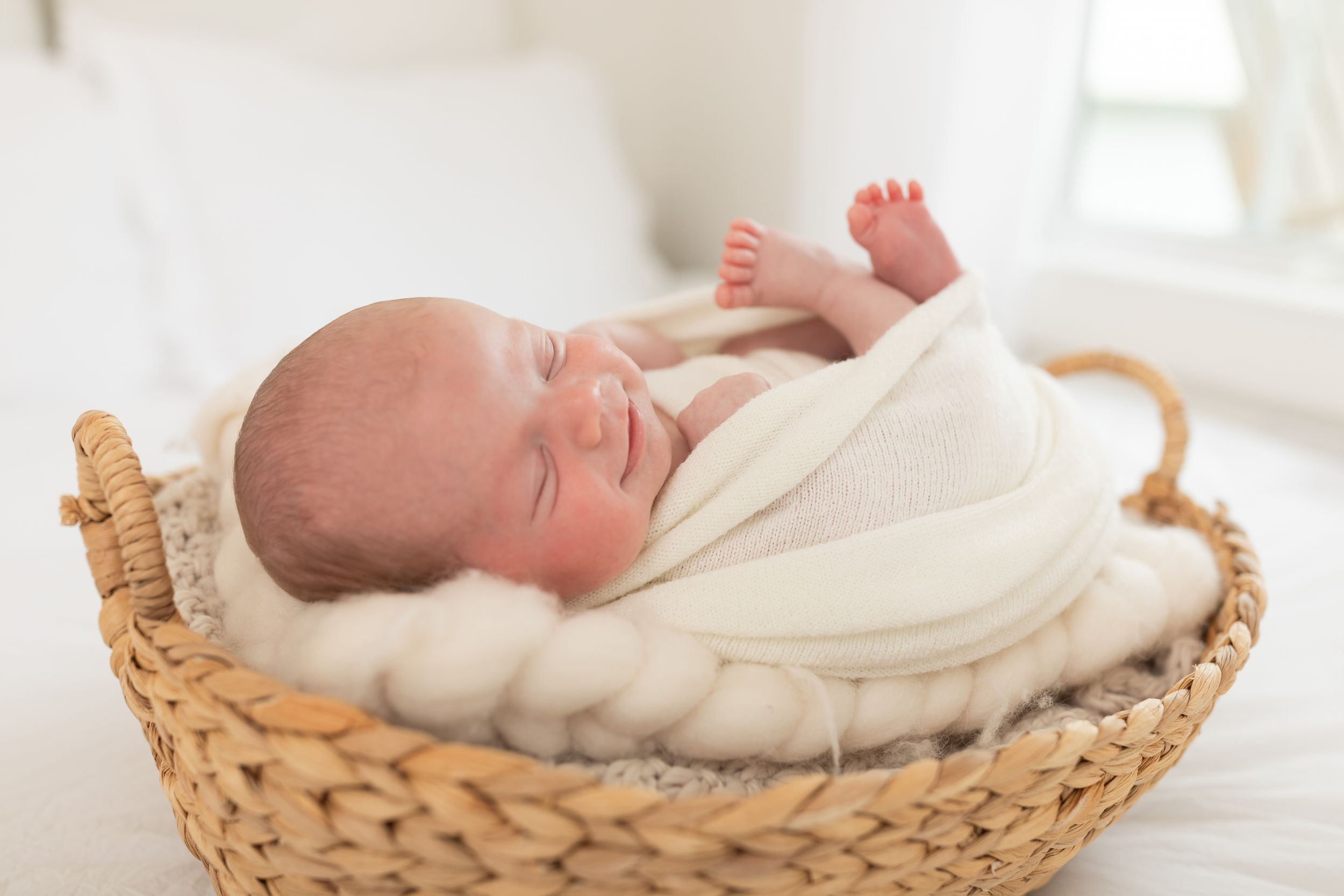 Newborn baby sleeping peacefully in a wicker basket with a soft white blanket in a bright, airy room.