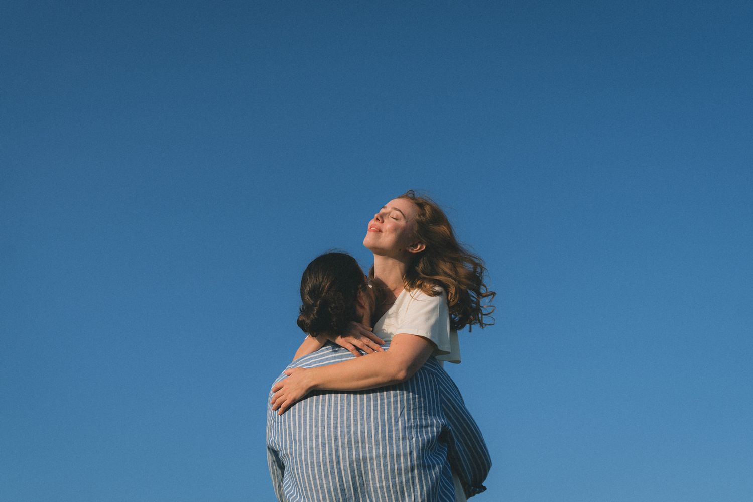 A couple embraces against a bright blue sky as the wind blows through their hair on a sunny day.