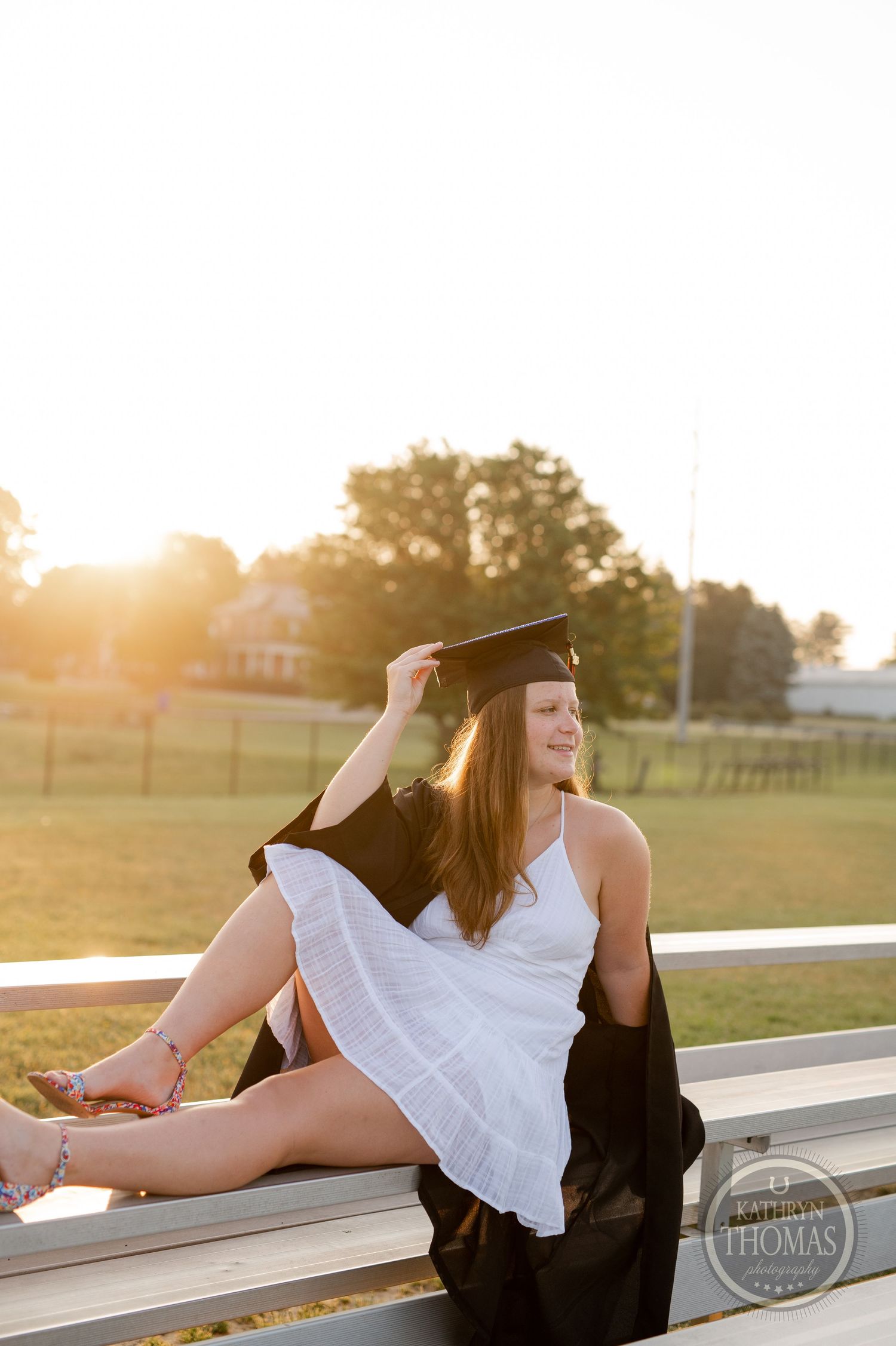 Graduate in white dress and cap posing on bleachers at sunset.
