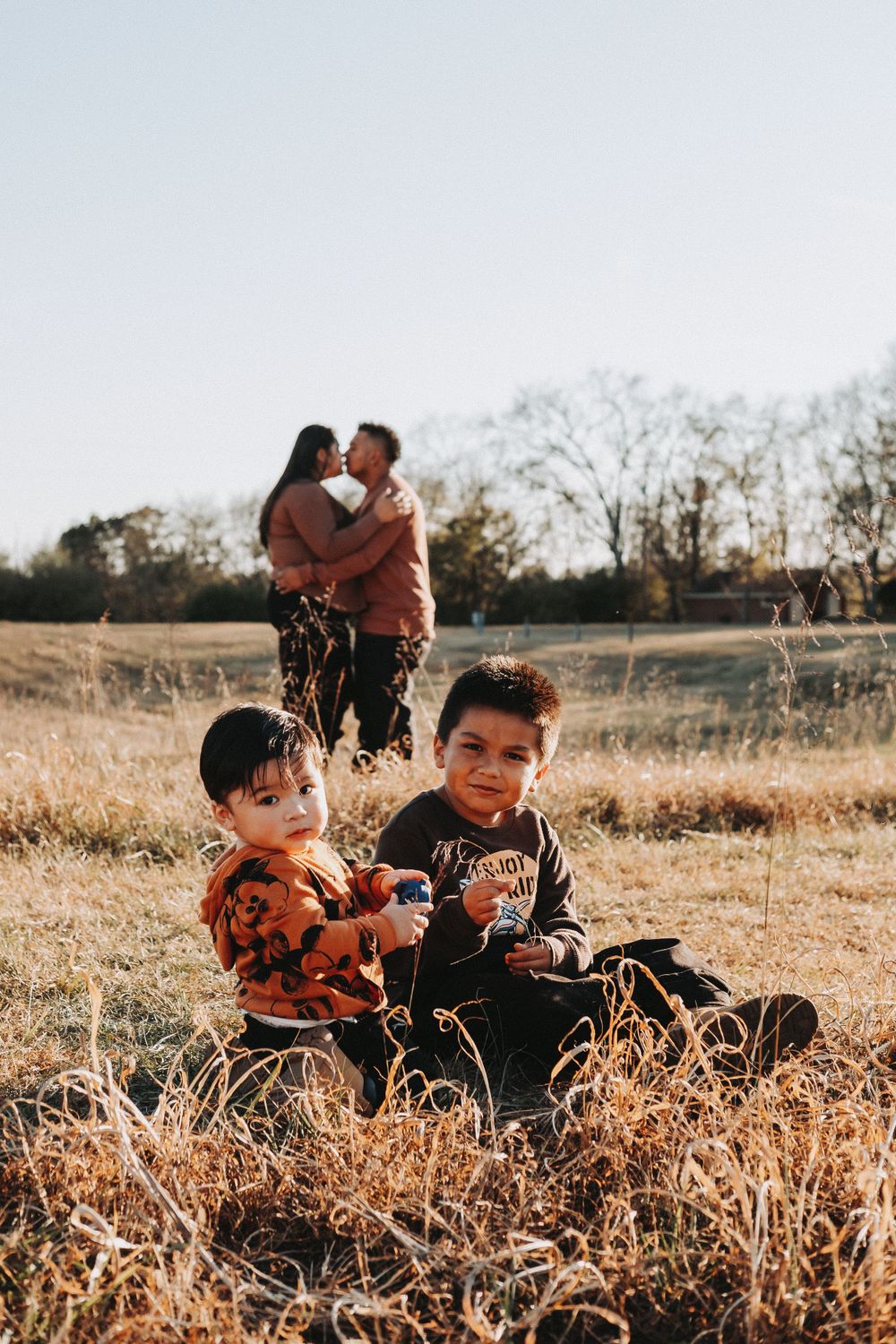 Children sit in golden field while adults embrace in background during sunset.